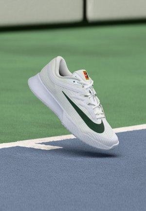 Scarpa da corsa Nike bianca con swoosh verde, che fluttua sopra un campo da tennis vicino alla linea di confine.