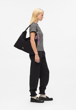 Black fabric shoulder bag with a curved shape, featuring a pink and purple accessory. Model wears a striped t-shirt and black joggers.