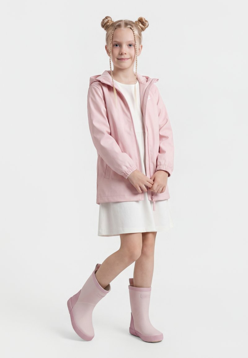 Jong meisje met gevlochten haar, een roze regenjas, een witte jurk en roze regenlaarzen, staande op één been.