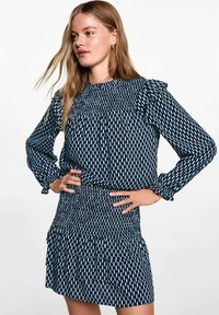 Marinblå klänning med vita lodräta mönster, med volangärmar och smockad midja, samt en flerlagerskjoldesign.