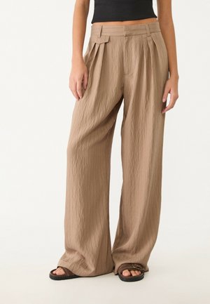 Wide-leg beige pants with pleats and a belted waist worn with black sandals and a black cropped top.