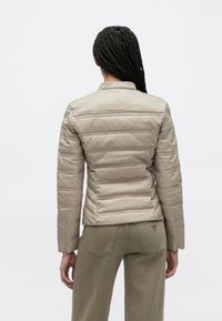 Smėlio spalvos dygsniuota striukė su aukštu apykakliu ir ilgomis rankovėmis, puošta subtiliais įspaustais raštais ir prigludusiu dizainu. Dėvima su alyvinių žalių spalvų kelnėmis.