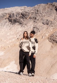 Twee modellen dragen beige sweatshirts met zwarte abstracte patronen en tekst. Ze combineren deze met zwarte broeken en sneakers, tegen een rotsachtige achtergrond.
