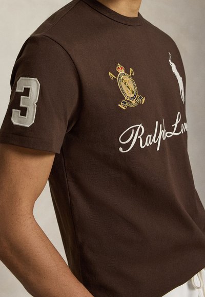 Άνδρας που φοράει καφέ πόλο Ralph Lauren με λευκό κεντημένο λογότυπο, χρυσό έμβλημα και μεγάλο λευκό αριθμό 3 στο μανίκι.