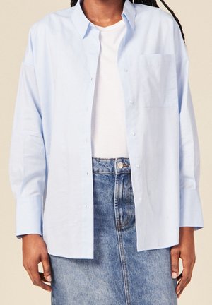 Personne portant une chemise bleu clair ouverte sur un top blanc assortie à une jupe en jean bleu taille haute, debout devant un fond neutre.