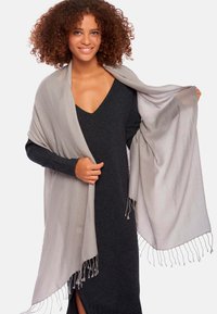 Châle gris clair en tissu doux, avec des bords frangés et une texture lisse, drapé sur un vêtement sombre et ajusté.