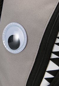 Grijze stof met een gestructureerd oppervlak, voorzien van een cirkelvormig plastic detail met een zwarte stip en een zwarte rits naast een witte, gemusterde accent. 
