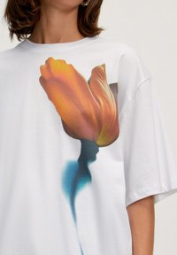 Weißes übergroßes T-Shirt mit einem lebhaften orangen Tulpenmotiv vor einem blauen Schatten. Aus weichem Baumwollmaterial mit lockerer Passform.