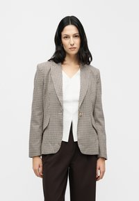 Blazer à carreaux en tissu beige et marron, doté d'une fermeture à un bouton, de revers à pointe et de deux poches avant. Porté sur un haut blanc.