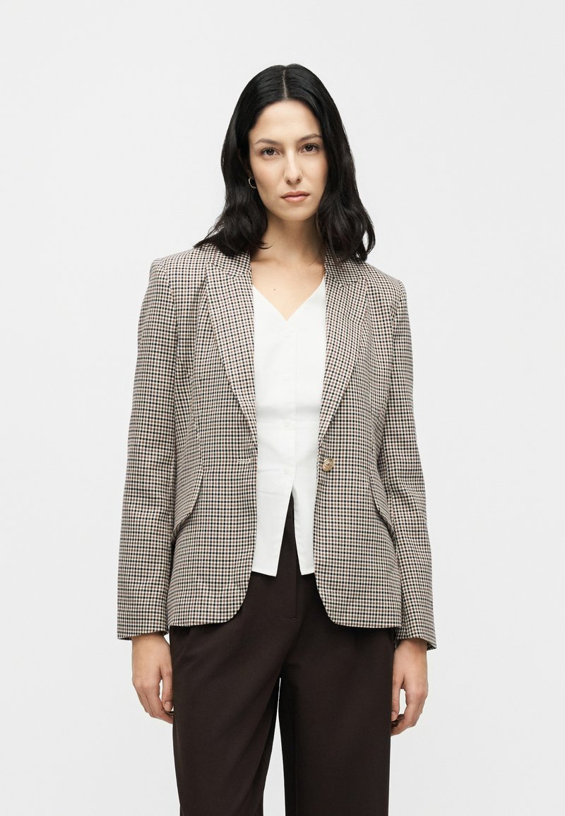 Blazer à carreaux en tissu beige et marron, doté d'une fermeture à un bouton, de revers à pointe et de deux poches avant. Porté sur un haut blanc.