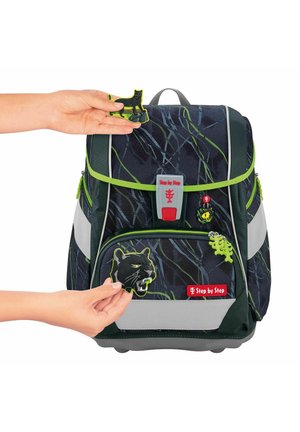 Der Rucksack verfügt über einen dunklen, gemusterten Stoff mit grünen Akzenten, mehrere mit Reißverschluss gesicherte Fächer und abnehmbare tierisch gestaltete Patches.