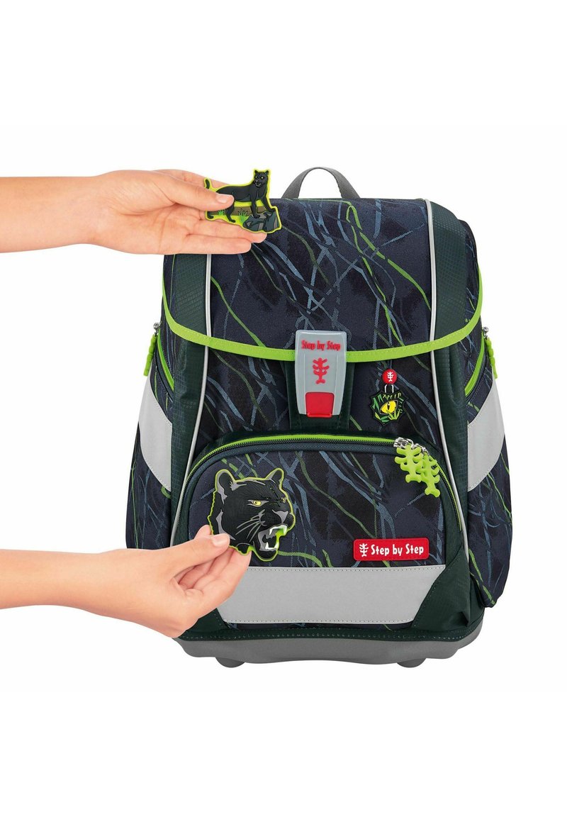 La mochila presenta una tela oscura con un patrón, acentos en verde, múltiples compartimentos con cremallera y parches temáticos de animales desmontables.