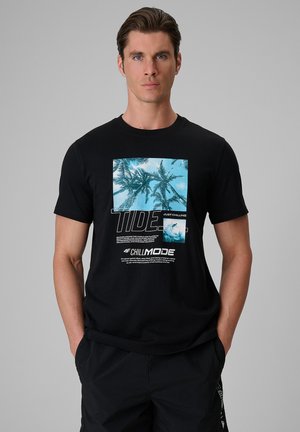 Mężczyzna ubrany w czarną koszulkę z niebieską grafiką palmy i tekstem "TIDE" oraz "CHILL MODE,", stoi z rękami w kieszeniach na jednolitym szarym tle.