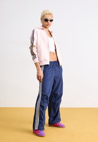 Rosa zip-up jacka med svarta ränder, vit crop top, blå träningsbyxor med vita ränder och lila sportskor. Slät textur.