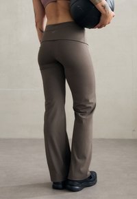 Brązowe spodnie sportowe z dopasowanym pasem i rozszerzanymi nogawkami, wykonane z gładkiego materiału. Logo Nike jest subtelnie umieszczone na pasie.