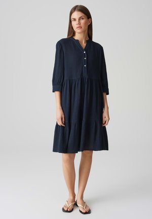 Navy blue cotton dress with a textured finish, three-quarter sleeves, buttoned neckline, and tiered skirt design, paired with flip-flops.