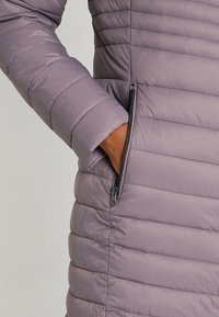 Lys lilla pufferjakke med horisontalt quiltet tekstur, der har en sidelomme med lynlås og tætsiddende ærmer. Glat, let materiale.