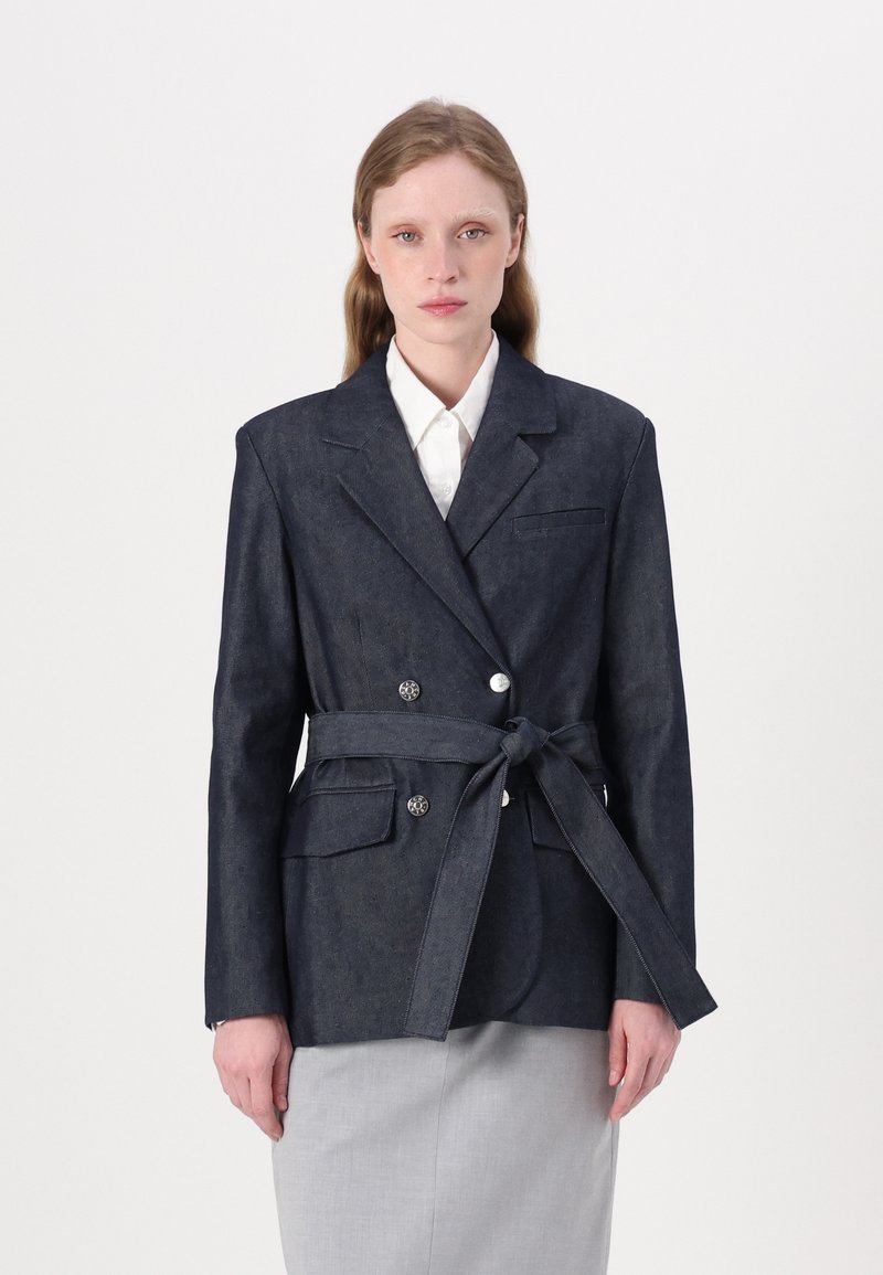 Sacou din denim închis, cu un design structurat, nasturi dublu rând, talie cu curea și două buzunare frontale. Purtați peste o cămașă albă.
