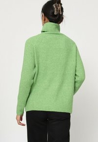 Femme avec des cheveux noirs dans une pince à cheveux en forme de griffes, portant un pull à col roulé côtelé vert et un pantalon noir, tournée dos à la caméra.