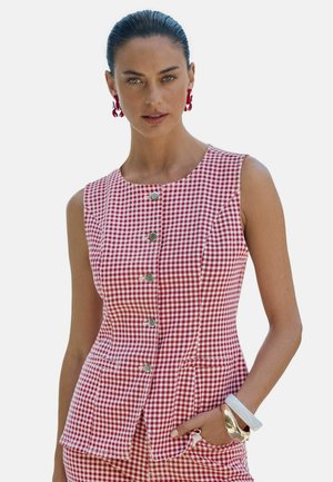 Kvinde med glatstrøget hår, iført en ærmeløs rød- og hvidternet knappet top og matchende bukser, accessoriseret med øreringe formet som hummere og armbånd.