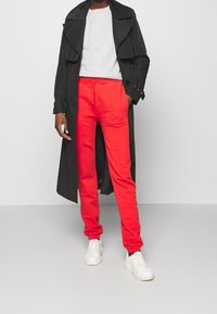 Black trench coat over a gray sweatshirt and red sweatpants with an elastic waistband. White sneakers complete the outfit.