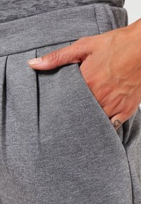 Szare materiałowe spodnie o gładkiej fakturze, z plisą z przodu i boczną kieszenią, ręka oparta na materiale.