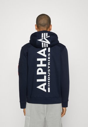 Tamnoplavi dukserica s kapuljačom koja ima veliki bijeli logo "ALPHA INDUSTRIES" na leđima, s kapuljačom na vezanje i rebrastim manžetama i donjim rubom.