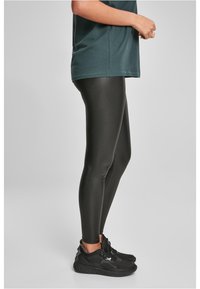 Leggings noirs taille haute avec une texture brillante, associés à un t-shirt vert foncé. Le pied du mannequin est chaussé d'une basket noire.