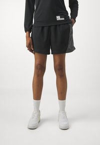 Pantalones cortos deportivos negros con textura de malla, con paneles laterales, usados con zapatillas deportivas blancas y calcetines de longitud media.