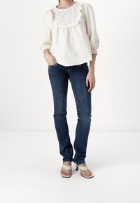 Blusa de algodón blanca con escote con volantes y detalles de ojales, combinada con vaqueros de pierna recta azul oscuro y sandalias de tacón beige.
