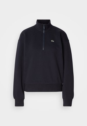 Marineblå hettegenser med høy krage og kvart glidelås, med lange ermer og ribbestrikket nederkant. Inkluderer en liten grønn logo på brystet.
