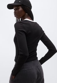 Sort langærmet top lagdelt over hvid skjorte, parret med mørkegrå jeans og sort kasket på person med fletninger, stående sideværts.