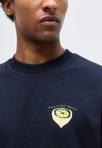 Κλείσιμο πλάνου ενός ατόμου που φοράει μπλε φούτερ με γραφικό λεμονιού και κείμενο «Marc O Polo Denim» στο στήθος.