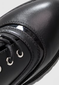 Sort lædersko med glansfulde detaljer, struktureret overdel, runde snørebånd og sølvøjer. Buede linjer skaber et strømlinet design.