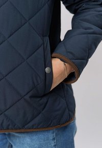 Tumši zils, krokots džinsa jaka ar dimanta raksta rakstu, brūnām kordūras detaļām uz aprocēm un snap pogu kabatas dekorā.