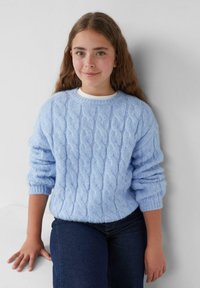Helles blaues Strickpullover mit Zopfmuster, rundem Ausschnitt und umgeschlagenen Ärmeln, getragen über einem weißen Oberteil und kombiniert mit dunklen Jeans.