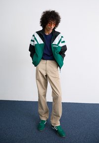 Green and black windbreaker with white stripes, navy t-shirt, beige trousers, and green shoes with white stripes. Curly hair and sunglasses.