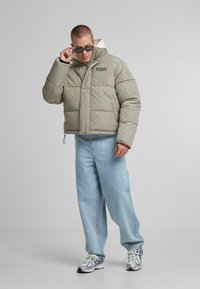 Polstret, lysegrå jakke med geometrisk mønster, frontlynlås, høj krave og logo-detalje. Bæres med løse blå jeans og sneakers.