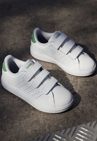 Baskets blanches avec trois bandes Velcro, accents verts sur le talon, détails perforés, en matériau synthétique lisse, semelle en caoutchouc texturé.