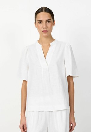 Žena s tmavými vlasmi zviazanými dozadu, oblečená v bielej blúzke s krátkym rukávom a golierom s výrezom do V a bielych nohaviciach na jednoduchom pozadí.