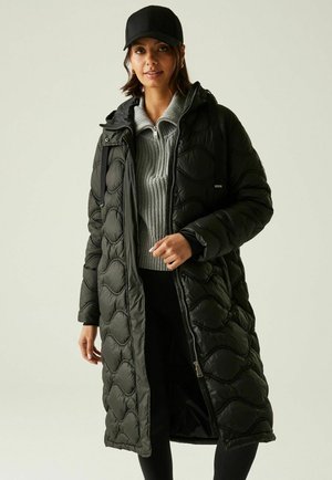 Femme portant un long manteau noir matelassé sur un pull gris à zip et une casquette noire, debout devant un fond clair uni.