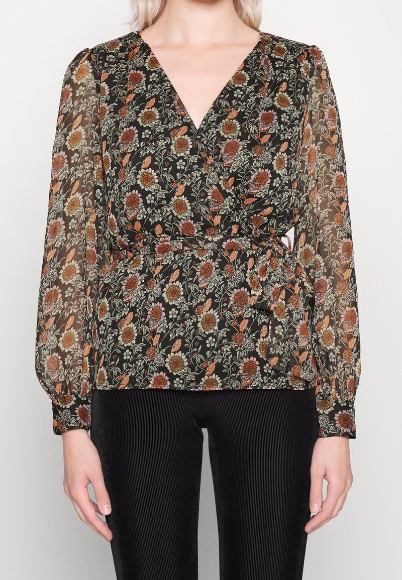 Femme portant un chemisier portefeuille noir avec de longues manches transparentes et un imprimé floral aux tons rouille, jaune et vert, associé à un pantalon noir.