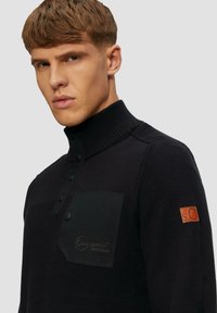 Schwarzer Strickpullover mit hohem Kragen, Knopfblende und einer strukturierten Tasche mit einem Logo. Lederakzent am Ärmel.