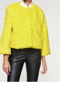 Faux fur yellow cropped jacket with a textured finish, wide sleeves, and a white ruffled collar. Black fitted leather pants complete the outfit.