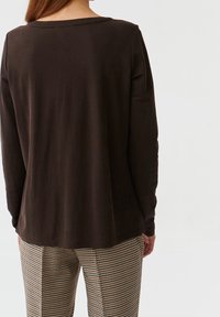 Haut marron à manches longues avec un col bateau, une texture lisse et un ourlet légèrement plus long à l'arrière, porté avec un pantalon à carreaux.