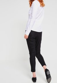 Vit långärmad skjorta ihop med svarta tajta byxor med dragkedjor i fållarna och svarta skor, sedd från baksidan mot en neutral bakgrund.