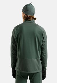 Dunkelgrüne Softshell-Jacke mit hohem Kragen, nahtlosem Schulterdesign und leicht strukturierter Oberfläche. Ausgestattet mit einer Reißverschlusstasche auf der Rückseite.