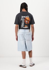 T-shirt graphique noir avec un design coloré de skull et de flammes au dos. Shorts en denim bleu clair et baskets blanches avec des accents noirs.