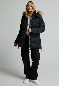 Giacca a vento nera imbottita con cappuccio foderato in pelliccia, chiusura con zip e patch con logo sulla manica; abbinata a pantaloni cargo neri e sneakers bianche.