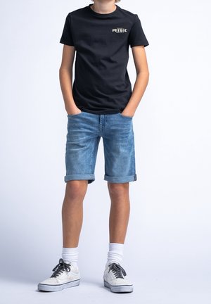 Camiseta de algodón negra con gráfico "PETROL", combinada con pantalones cortos de mezclilla azul claro con dobladillo enrollado. Calcetines blancos y zapatillas blancas.